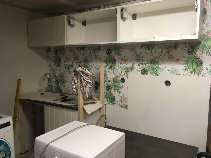 Överskåp installerade i kök med tropisk tapet, oskarpa verktyg på bänkskiva, diskho och en vit tvättmaskin.