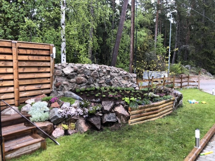 Nybyggd stenmur i en trädgård med blommande växter och gräsmatta, intill en trägrind och staket.