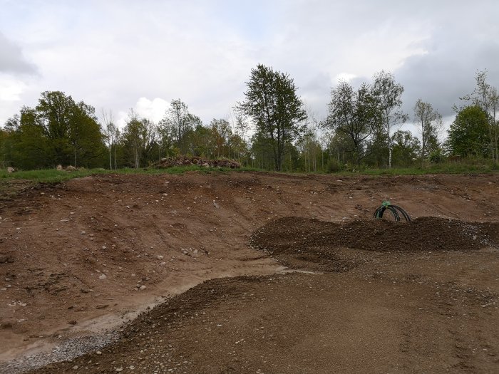 Obebyggd tomt med sluttning och stora högar av grus, mot bakgrund av lövträd och himmel.