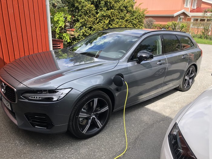 En grå laddhybridbil parkerad och laddad framför ett hus med röd vägg.