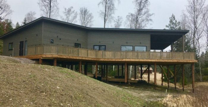 Hus byggt på höga stolpar över en sluttning med träterrass och brant backe i förgrunden.