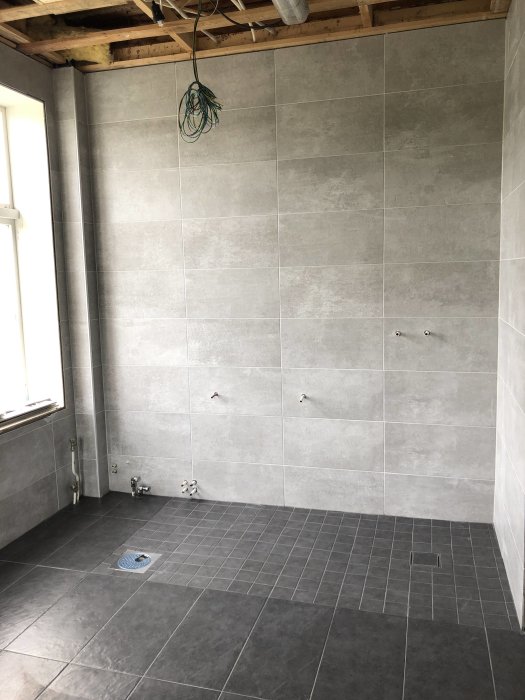Badrum under renovering med stora klinker på vägg och mindre klinker på golv, synliga rör och två golvbrunnar.