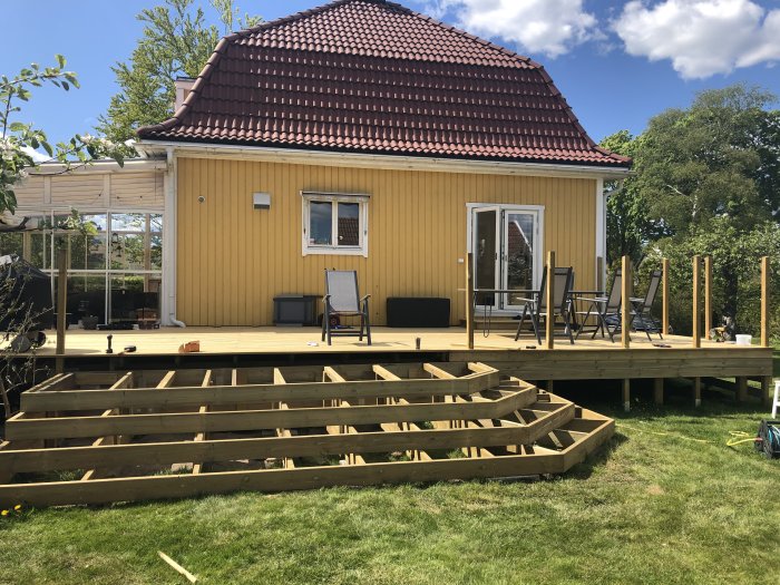 Pågående bygge av en träaltan vid ett gult hus med klarblå himmel i bakgrunden.