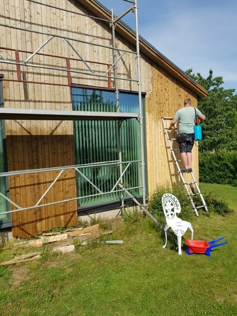 Person på stege arbetar med träfasad på hus, byggmaterial och verktyg synliga på gräsmattan.