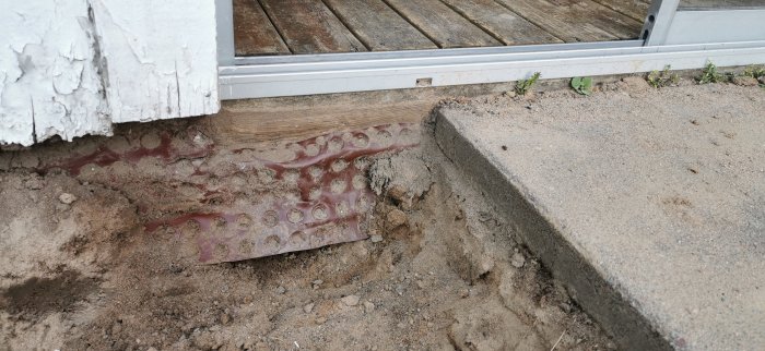 Vinröd fuktspärrsfolie som skyddar en träplanka vid ett tröskelspår vid en altandörr, med sand och betong intill.