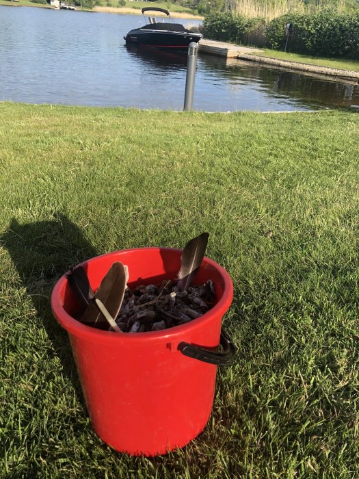 Röd hink med gödsel på gräs framför en flod med en båt i bakgrunden.