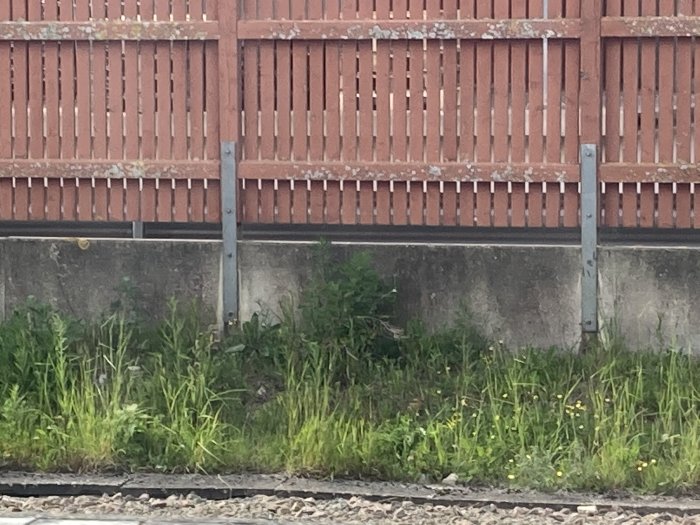 Ett högt plank längs en tågstation med metallstöd på en betongmur, omgiven av grönska.