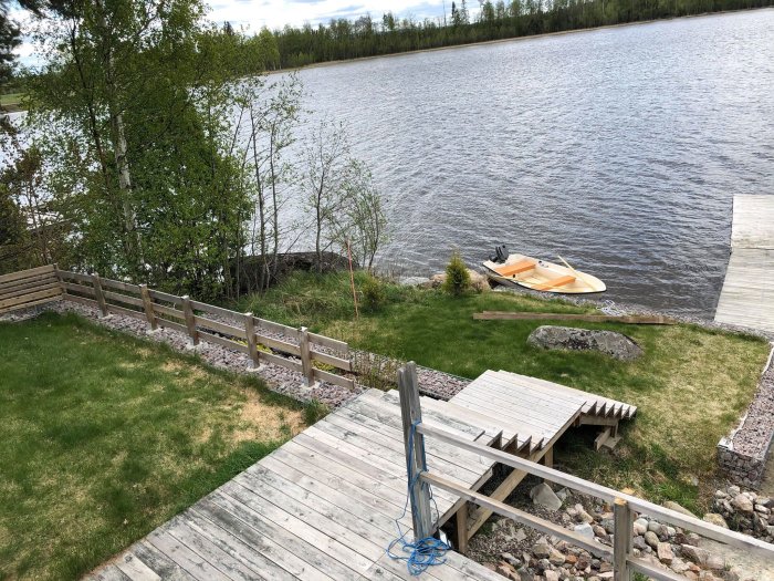 Trädgård i etage vid sjö med staket, stenkantningar och en brygga med båt.