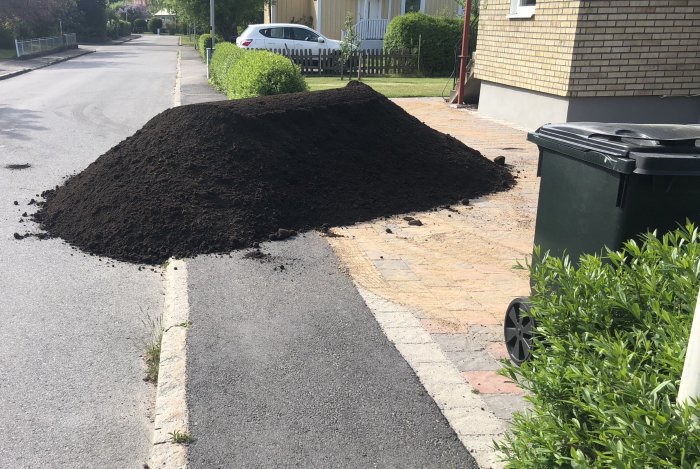 Stor hög med mörk påsjord på en uppfart bredvid gröna soptunnor och en bil i bakgrunden.