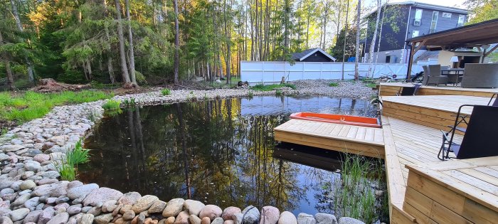 Trädgårdsdamm omgiven av stenar med trädäck och brygga, röd båt på vattnet och hus i bakgrunden.