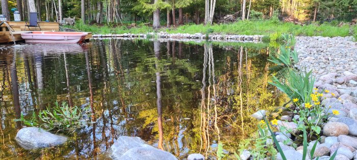 Naturtomt vid vattenkanten med en rosa båt, stenmur och vegetation reflekteras i lugnt vatten.