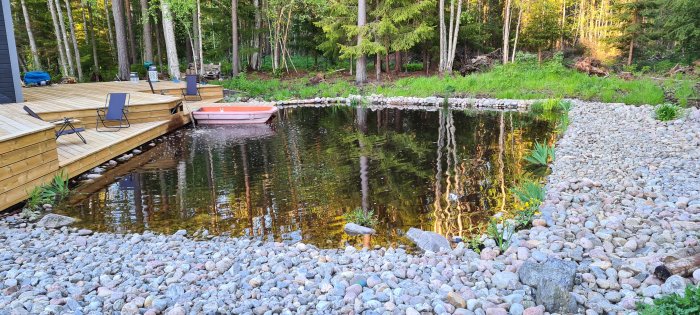 Trädäck vid en skogssjö med en rosa roddbåt och strandkant med runda stenar.