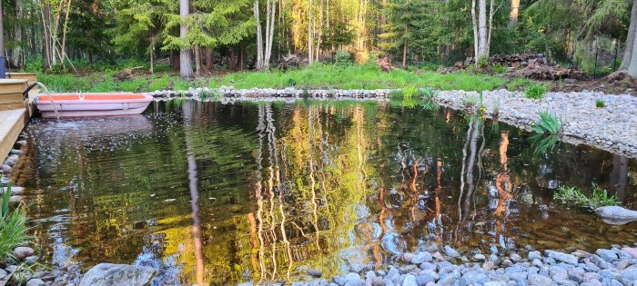 Skymning vid en lugn skogssjö med spegelblank yta, en orange roddbåt vid bryggan och stenlagd strandkant.