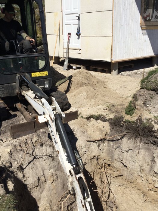 En grävmaskin av märket Bob-Cat E16 utför markarbete vid grunden till en byggnad, med synliga gamla plintar och skalad jord.