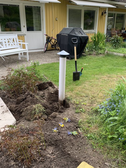 Vit ventilationsrör sticker upp i en rabatt med spade och uppgrävd jord vid en gul husvägg.