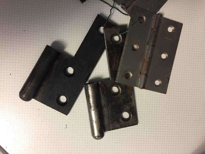 Två behandlade gångjärn med hård svart yta jämförs med ett blankt obehandlat gångjärn på en vit bakgrund.