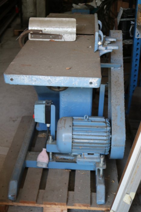 En blå stationär träbearbetningsmaskin med motor och rullbord i en verkstad.