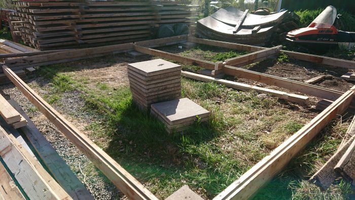 Grundläggning av bastustuga med virke och betongplintar, verktyg och byggmaterial syns i grönområde.