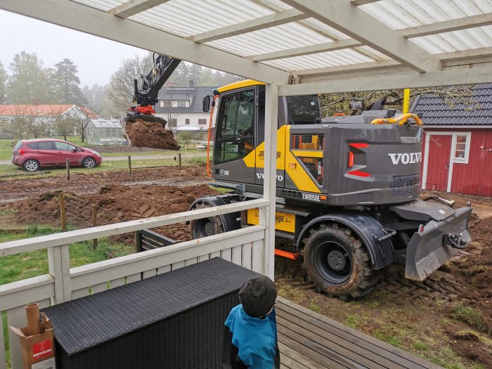 Gul grävmaskin flyttar jord vid förberedelse av avlopp och el till ett garagebygge.