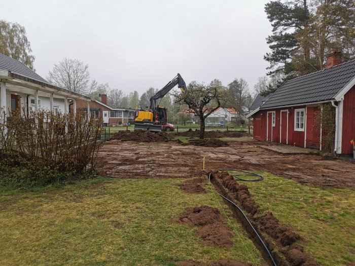 Grävmaskin som förbereder avlopp och EL/vatten till ett garagebygge i ett upprivet trädgårdsområde.