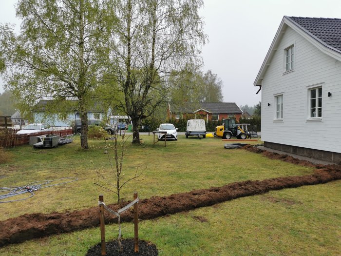 Grävd ränna för avlopp och el i trädgård nära vit husfasad, med grävmaskin och material i bakgrunden.