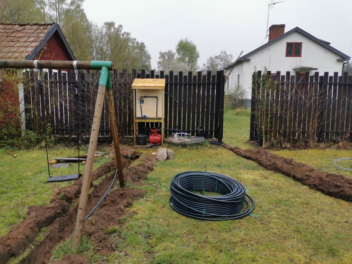 Grävd ränna i trädgård för avlopp och el med slangrulle, stegar och en byggnad i bakgrunden.