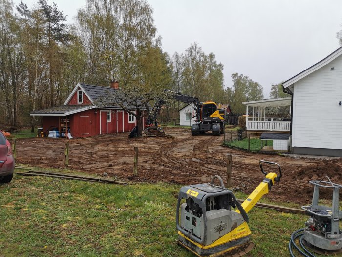 Byggarbetsplats med uppgrävd mark och grävmaskin vid ett rött hus och ett vitt garage.