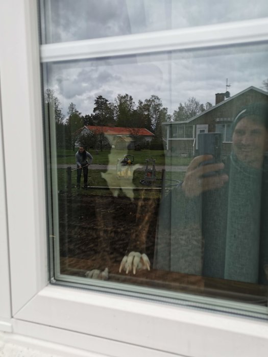 Fotografering genom fönster av en person utomhus som förbereder avlopp och EL/vatten vid ett garageprojekt.