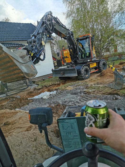 Grävmaskin lyfter grävskopa nära ett hus med grävt område i förgrunden, person håller en dryckesburk.