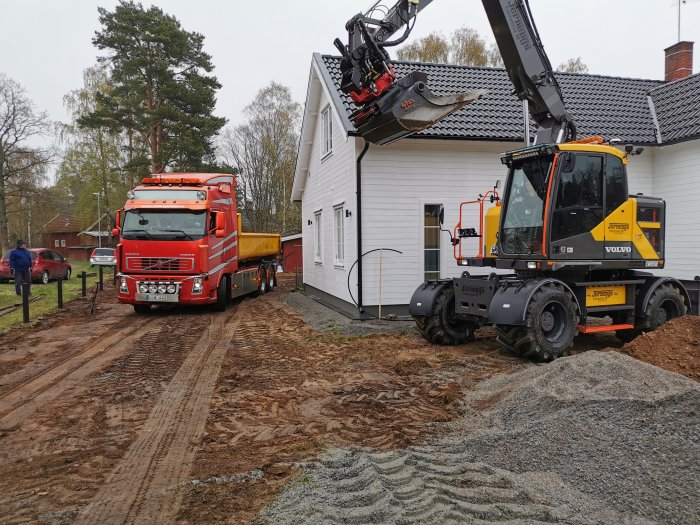 Grävmaskin och lastbil vid garagebygge med synliga avlopps- och el/vattenförberedelser.