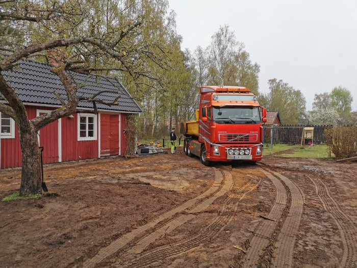 Lastbil vid pågående garagebygge med grävd mark och avloppsarbete i en trädgård.