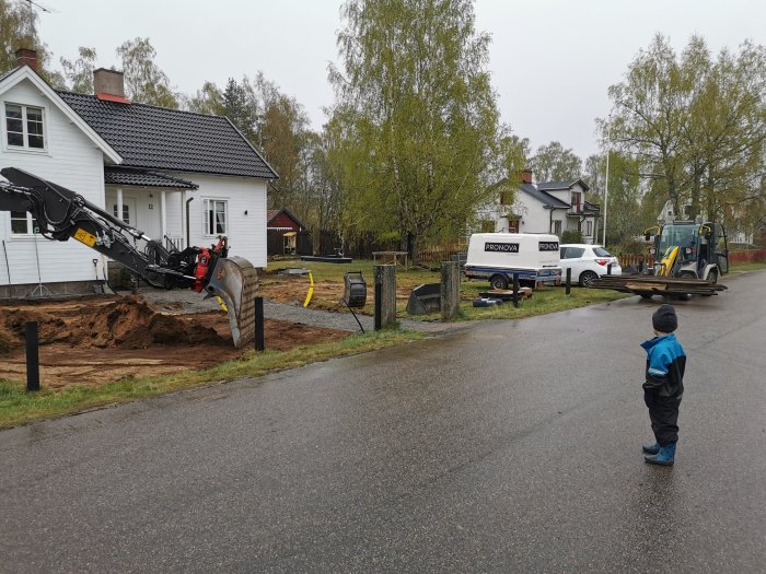 Grävmaskin gräver för avlopp vid vit villa, barn tittar på, hus och bilar i bakgrunden.