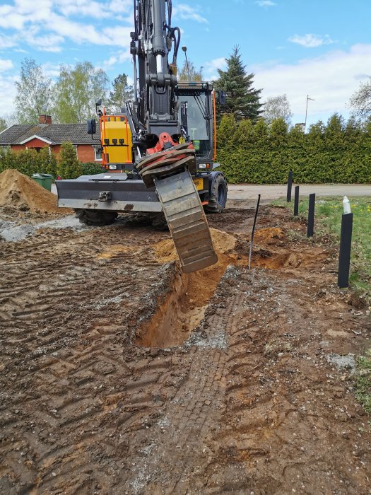 Grävmaskin förbereder avlopp och EL/vattenledningar vid garagebygge, arbetsområde med uppkört mark och stolpar.