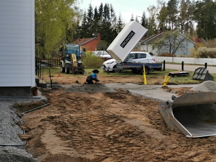 Grävplats för avlopp och el/vatten vid garagebygge, person arbetar, grävmaskin och utspridda rör i förgrunden.