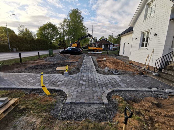 Pågående arbete med att lägga marksten i ett U-format mönster framför ett hus, med grävmaskin i bakgrunden.