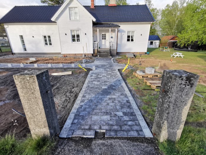 Nyinstallerad grå och antracit marksten leder fram till ett vitt hus med blått tak, omgivet av pågående trädgårdsarbete.