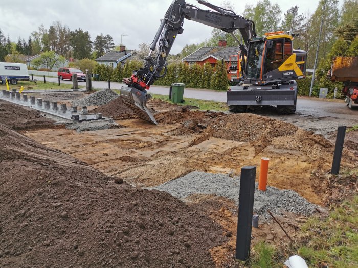 Grävmaskin som förbereder mark för stenkista, med grus och marksten vid en parkering.