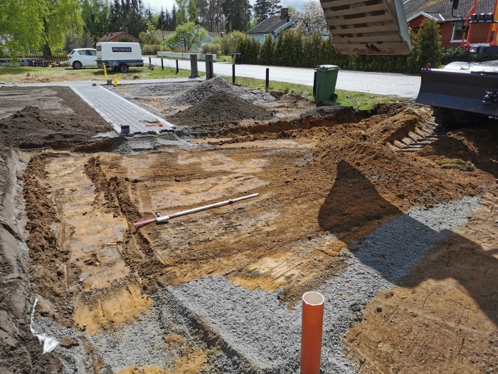 Byggplats med grus, högar av jord, påbörjad stenläggning och parkerade fordon i bakgrunden.