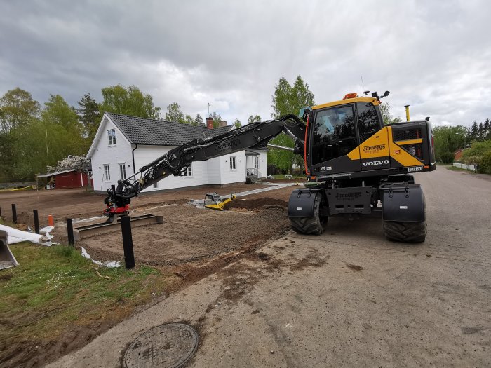 Grävmaskin i arbete framför ett vitt hus, förbereder mark för anläggning av parkering.