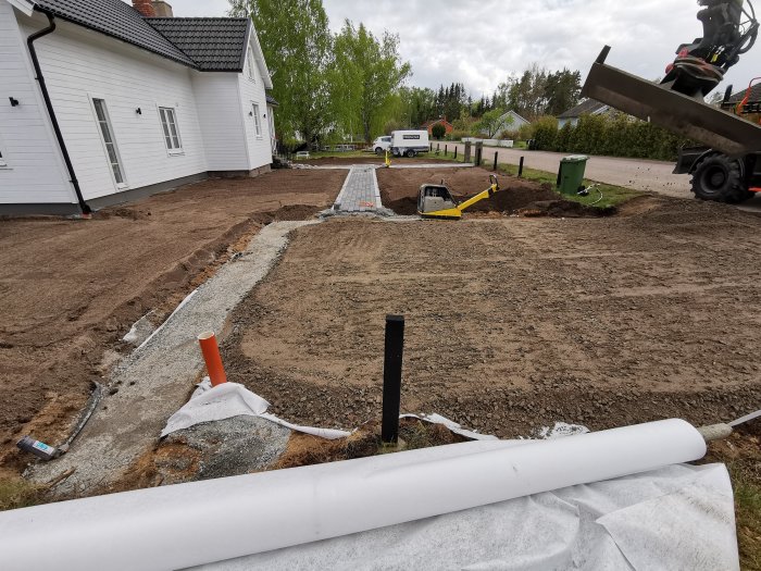 Område under konstruktion med grävmaskin, stenkista och preparerad mark inför markstensläggning vid ett hus.