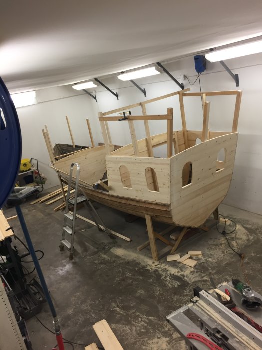 En träbåt under konstruktion i ett garage med verktyg och byggmaterial runtomkring.
