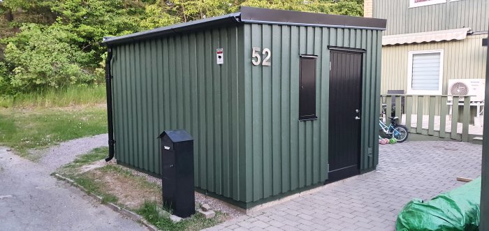 Nybyggd grönmålad förrådsbyggnad med nummer 52, svart dörr och brevlåda, intill ett hus.