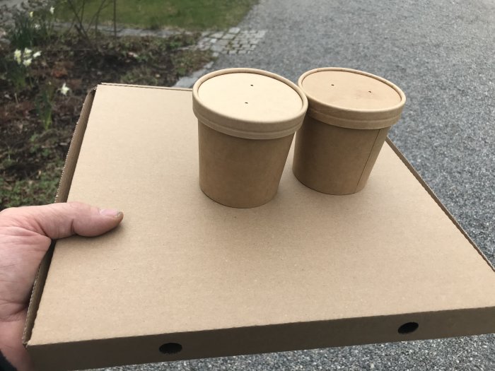 Hand som håller en brun papperspizzakartong och två bruna pappmuggar med lock utomhus.