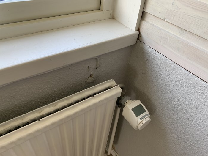 Radiator med termostat nära fönster i en fastighet, viss slitage synlig.