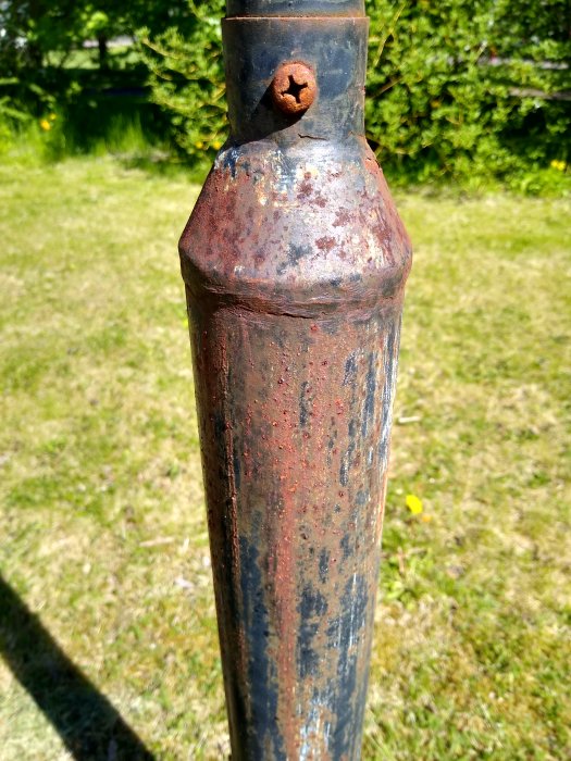 Närbild av en gammal metallstolpe med rost och färgflagning, belägen i en trädgård.