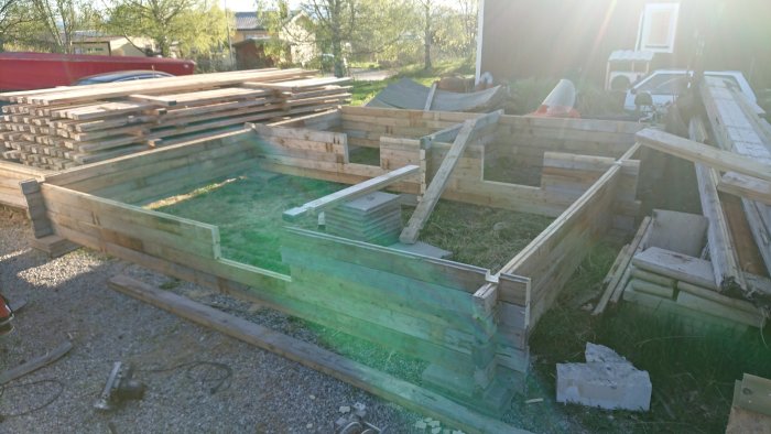 Halvfärdigt träbyggnadsprojekt utomhus med trähögar och en bränd cirkelsåg på marken.
