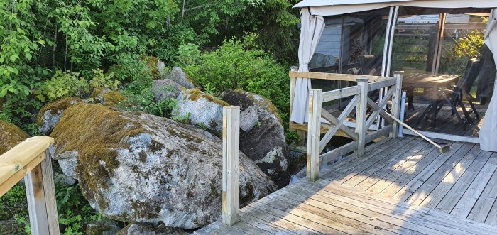 Pågående utbyggnad av en trädäck altan nära stora stenar med skog i bakgrunden och en sopborste på trallen.