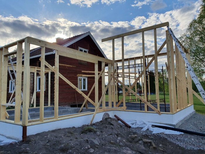 Byggprojekt med pågående konstruktion av en trästomme på en gjuten grundmur, bredvid en äldre träbyggnad.