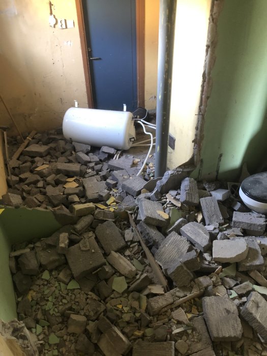 Rivningsavfall och tegelstenar på ett golv inne i ett delvis raserat rum med en omkullvält vit varmvattenberedare.