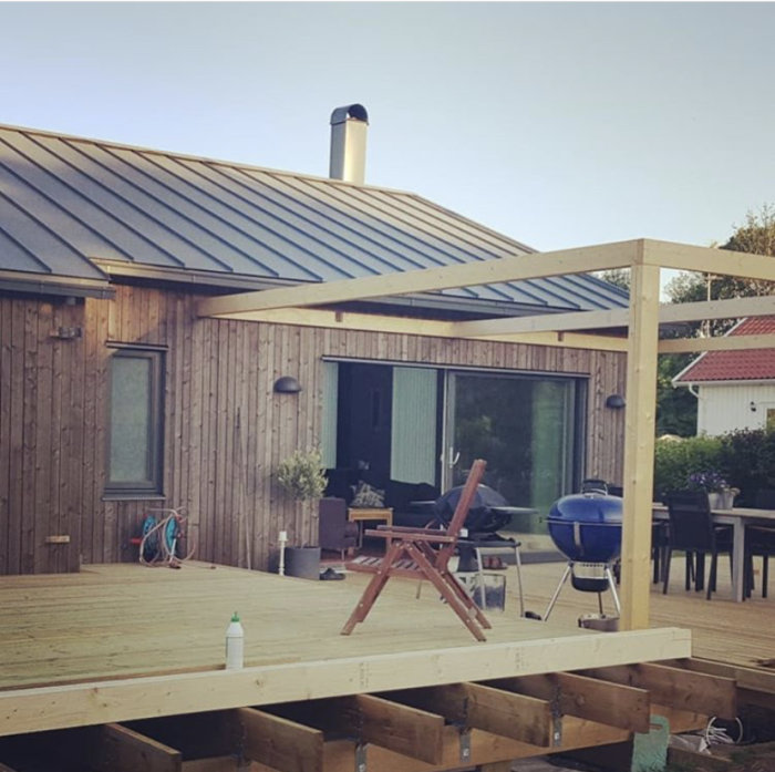 Nybyggd trädäck och pergola vid ett trähus med en del trädgårdsmöbler och en grill.